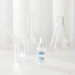 Dr. Belmeur Spot Care Ampoule 22ml – beruhigendes Serum mit Panthenol, Beta-Glucan, Chaga-Pilz und Teebaumöl gegen Unreinheiten und Rötungen. Abbildung Produkt mit Reagenzgläser im Hintergrund.